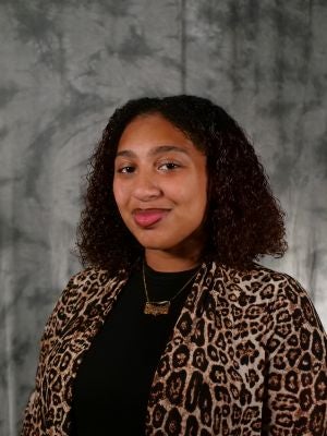 College-aged female student, dark curly shoulder length hair. Black shirt, cheetah print cardigan. Gold necklace. Smiling. Dark eyes.