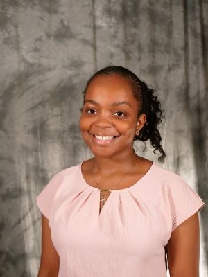 College-aged female student, dark hair, fulani braids pulled back into pony-tail, hair is wavy, dark eyes, smiling with teeth, pink blouse, gold necklace, diamond earrings