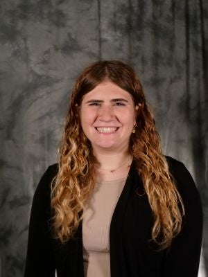 College-aged female student. Ombre colored, long wavy hair, brown on top of head blended to a dark blonde hair, dark eyes, smiling with teeth, tan shirt, black blazer, gold necklace with a small pink heart, gold earrings
