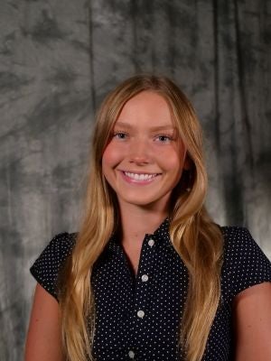 College-aged female student. Long blonde hair. Blue eyes. Smiling with teeth. Black and white polka dot short sleeve blouse with buttons.