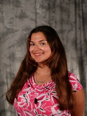 College-aged female student, medium brown long hair. Dark eyes. Smiling with teeth. Pink blouse with a floral pattern.