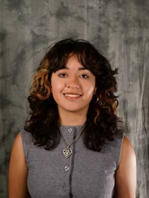 College-aged female student, dark hair with gold highlights, wavy hair with bangs, dark eyes, smiling with teeth, grey sleeveless button up blouse, silver heart-shaped necklace, silver hoop earrings