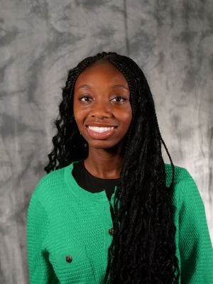 College-aged female student. Dark, long, boho braids. Dark eyes. Smiling with teeth. Black shirt with green long-sleeve knit-style sweater with brown buttons.