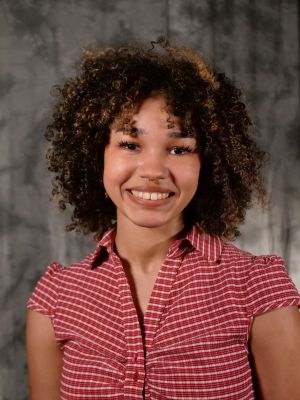 College-aged female student, dark hair with blonde highlights, curly, dark eyes, septum piercing, smiling with teeth, red and white shortsleeve button up collared blouse
