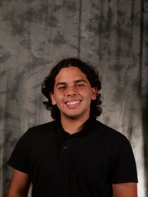 College-aged male student. Dark wavy hair. Medium length. Dark eyes. Smiling with teeth. Black polo.