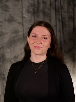 College-aged female student. Dark long hair. Light eyes. Smiling. Black blouse, black blazer. Gold necklace with charm. Gold earrings.