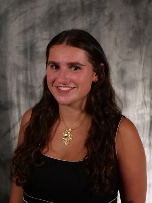 College-aged female student. Dark, long, wavy hair. Dark eyes. Smiling with teeth. Black sleeveless dress with white outlining. Gold necklace with large gold charm with diamonds and red in middle.