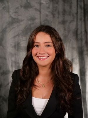 College-aged female student. Brown wavy long hair. Dark eyes. Smiling with teeth. White shirt covered with black blazer. Gold necklace with charm. Earrings.