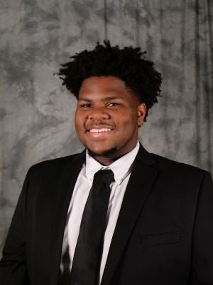 College-age male student. Black fro. Facial hair. Dark eyes. White button up with black suite jacket and black tie. Smiling with teeth. Earring.