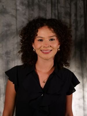 College-aged female student. Dark curly hair. Medium length hair. Dark eyes. Smiling with teeth. Gold necklace with 
