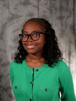 College-aged female student. Dark hair with fulani hair style. Dark eyes. Dark framed glasses. Smiling with teeth. Green long sleeve blouse with brown buttons.