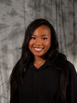 College-aged female student. Dark long hair. Dark eyes. Smiling with teeth. Black button up blouse. Gold necklace with multiple diamonds on chain.