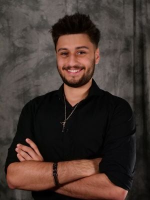 College-age male student, dark hair, dark eyes, facial hair, smiling with teeth, wearing black button up shirt, and a silver chain, with his arms crossed