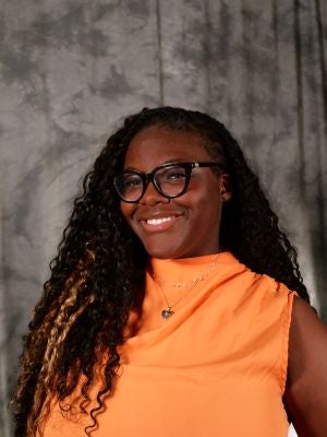 College-aged female student. Boho braids. Smiling with teeth. Silver necklace with heart charm. Black glasses. Orange blouse.