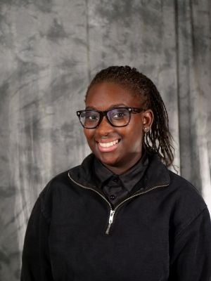 College-aged female student, dark hair, twists, in a pony tail, dark eyes, wearing black frame glasses, smiling with teeth, black button up, black quarter-zip, silver earrings