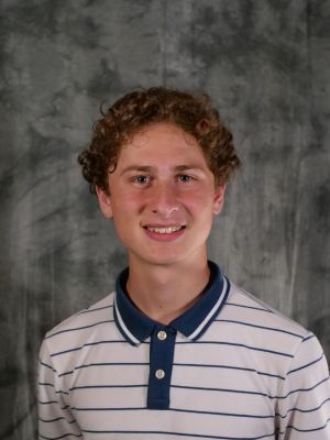College-age male student, reddish-brown curly short hair, light eyes, smiling with teeth, white polo with small blue horizontal blue stripes