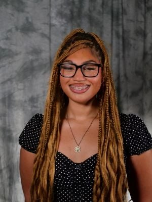 College-aged female student. Bronde long hair, french curl braids, loosened at the ends. Dark eyes. Black framed glasses. Smiling with teeth. Braces on teeth. Black and white polka dot short sleeve blouse with buttons. Gold necklace