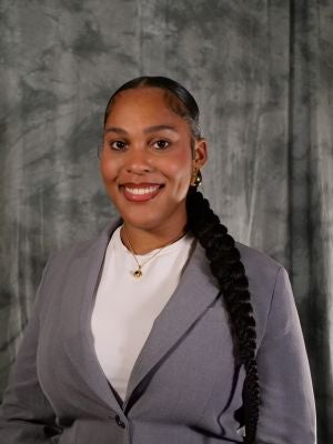 College-aged female student, dark hair in a braid pony tail that is lying down her shoulder and front, dark eyes, smiling with teeth, white shirt covered with grey blazer, gold necklace, gold earrings