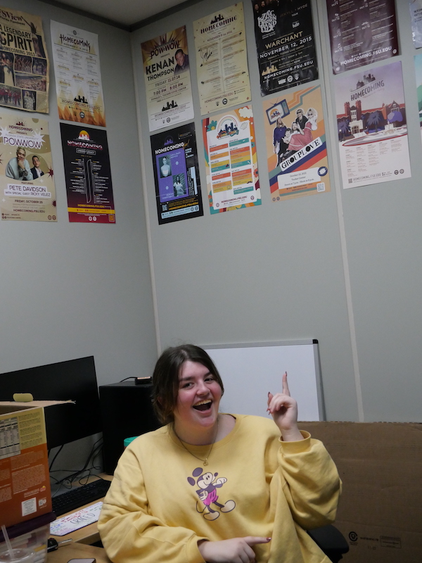 Corren Mahone points to a wall of Homecoming flyers behind her.
