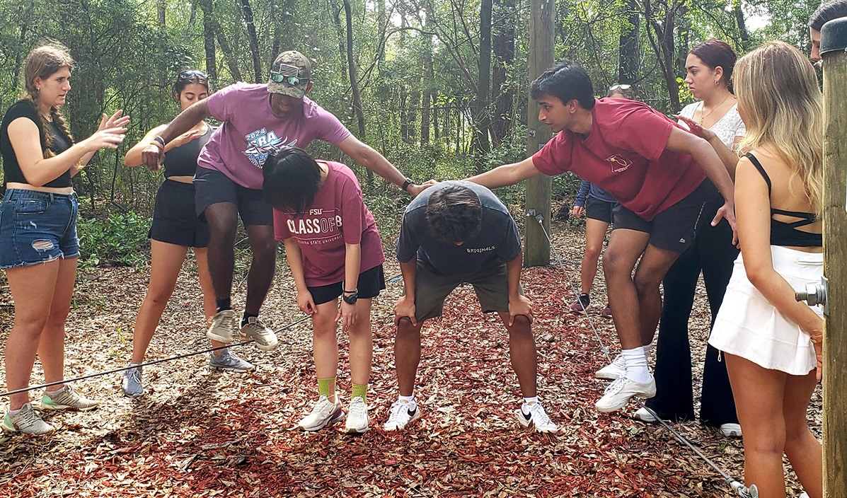 "Students gather around two peers working together to balance on low wires on the FSU challenge course"