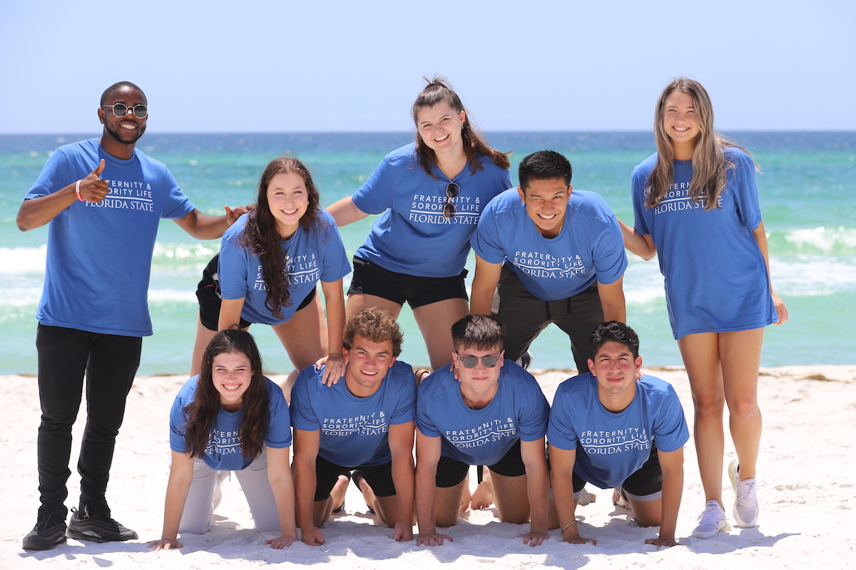 "A group of students pose in a pyramid on the beach with the water behind them."