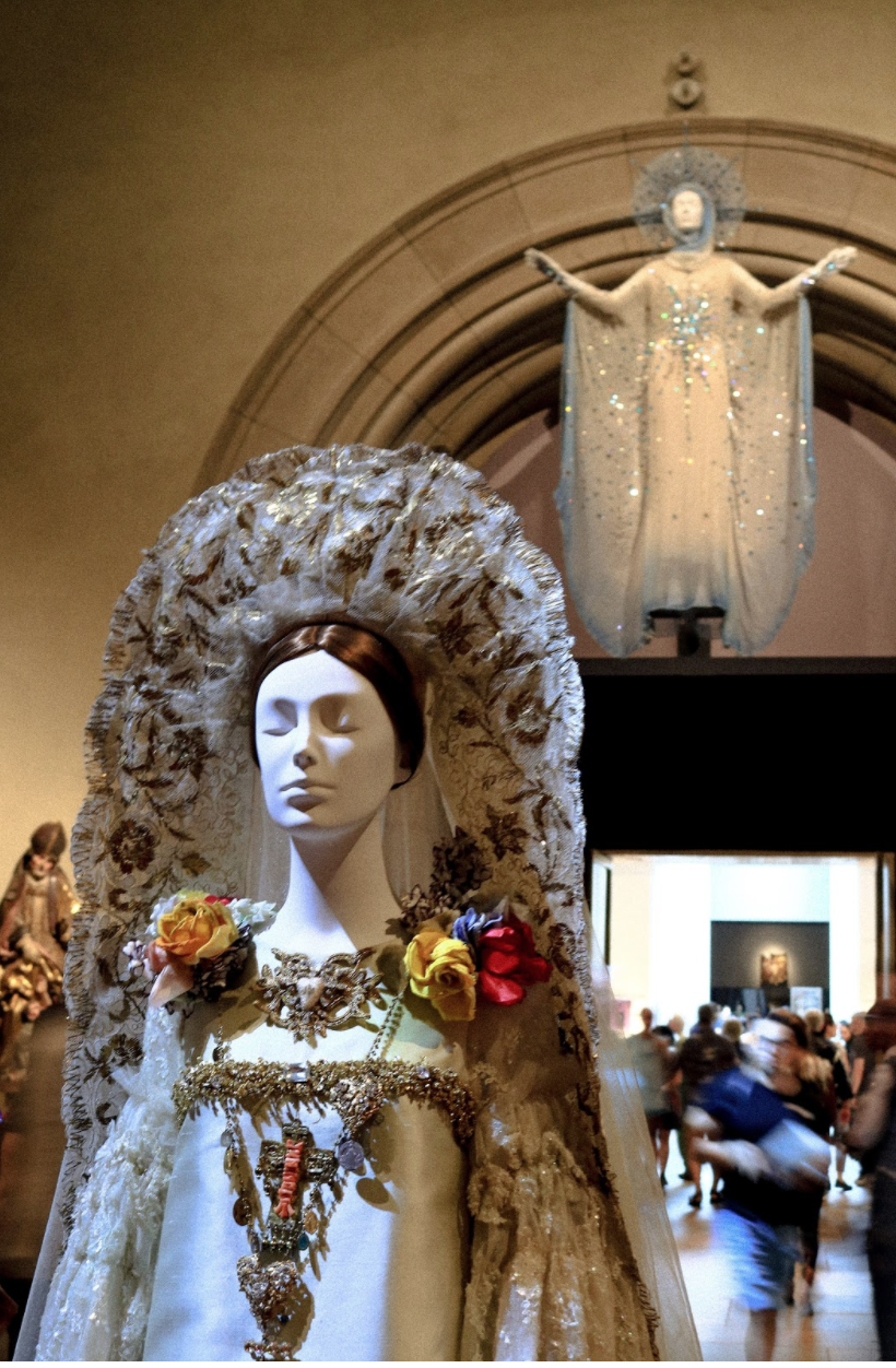 Museum setting with a mannequin in the foreground and mid-ground, each dressed in white robes. Crowd of museum goers in the background.