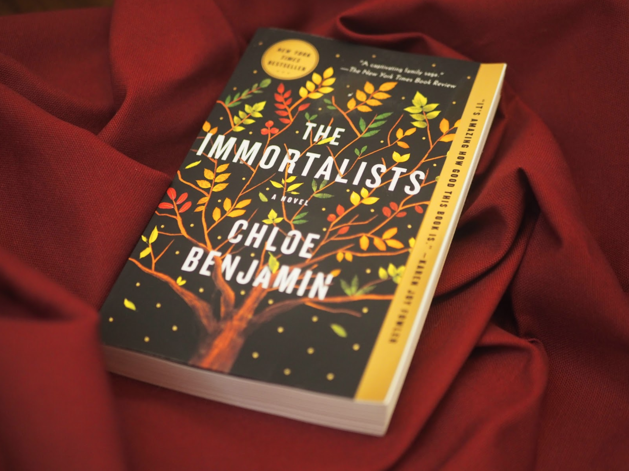 Paper copy of the book, "The Immortalists," by Chloe Benjamin. The novel cover has a black background with a tree of life, with leaves that have the appearance of fall-colored ferns.