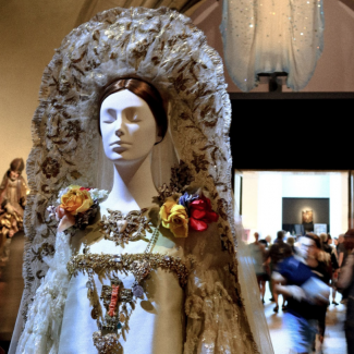 Museum setting with a mannequin in the foreground and mid-ground, each dressed in white robes. Crowd of museum goers in the background.
